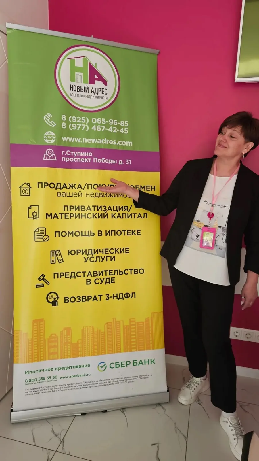 Татьяна Савельева, агент по недвижимости, у баннера агентства со списком услуг